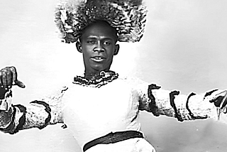 Black and white photograph of a Black Man in a white dress with dark ribbons on the sleeves and a fabulous headpiece that is really hard to describe.