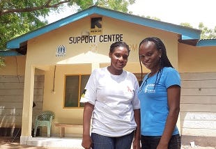 Two women — Phloviance Osumba and Dorcas Onkwani — stand in front of a building labeled “IRC Support Centre.”
