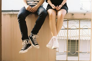 A man and woman sitting on a wall. Only their legs are visible, with both wearing Converse sneakers.