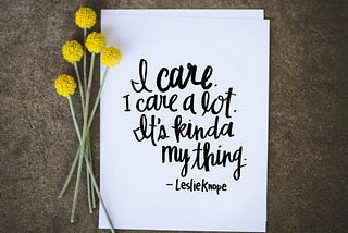 Yellow wildflowers and a stone surface nest to a sign reading, “I care. I care a lot. It’s kinda my thing. — Leslie Knope”