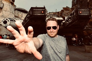 Justin Cox wearing sunglasses, holding his hand toward the camera as if channeling the Force. In the background in the Millennium Falcon.