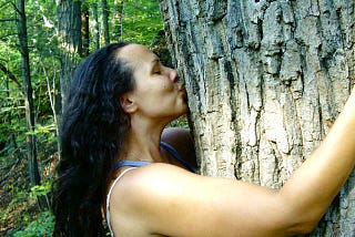Profile of a woman with her arms wrapped around a tree while kissing the tree. Her eyes are closed.