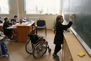 Young girl with disability in a classroom. She is writing on the blackboard, standing and the wheelchair behind her.