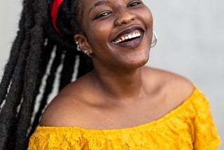 Smiling black woman wearing a bright yellow blouse. She is also wearing dreadlocks.