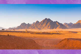 A panoramic view of a jagged mountain range from the point of view of a mile away in an orange-dirt desert.