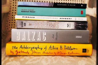A stack of books about Paris include Giovanni’s Room by James Baldwin, Monsieur Proust by Céleste Albaret, Illuminations by Arthur Rimbaud, Parisians by Graham Robb, Lost Illusions by Honoré de Balzac, and The Autobiography of Alice B. Toklas by Gertrude Stein.