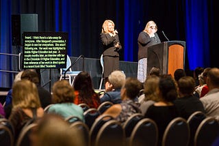 A scene from a conference presentation showing the audience sitting, a speaker presenting, and two accessibility supports: a sign language interpreter and a real-time captioning screen.