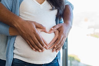 A close-up image of a pregnant woman holding her hands in the shape of a heart on her stomach along with her partner’s hands.