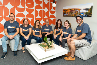 Seven members of the Architectural Designs team pose in an office with a bright orange and write wall. They are wearing blue shirts that read Bueno to honor AD alumnus Ryan Bard, who is pursuing his lifelong dream of becoming a commercial pilot.