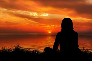 A silhouette of a woman facing the setting sun and meditating on a bank of a river