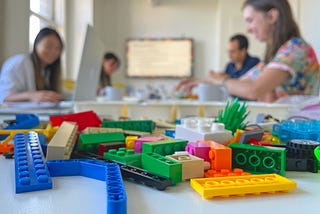A pile of colourful assorted LEGO bricks scattered on a table in the foreground with four workshop participants blurred in the background building their own models in a boardroom