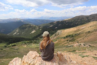 The back of a woman sitting on a rock staring out at a mountain range