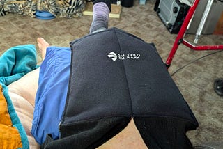 A person is lying down with their leg elevated and wrapped in a black therapy device, indicated by the label “SM FOLD THERAPY.” Surrounding them are various household items, including a blue blanket and a red walker.