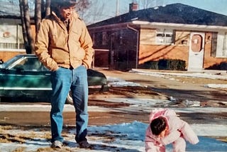 My father, a young brown-skinned man with black hair and glasses, is looking down and smiling at me as a toddler bending down to touch the bits of snow left on the ground. He is wearing a brown jacket and blue jeans and has his hands in his pockets. I am wearing a pink puffy snowsuit. We are in the front yard of a suburban neighborhood with brick ranch homes.