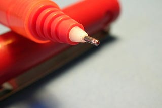 Picture shows a close up of a red pen with the cap removed. The pen is resting on the cap such that the point is raised.