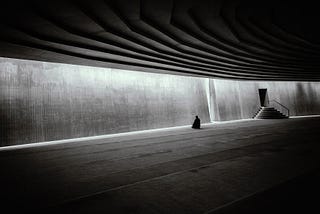 Black and white. A figure in the distance of a concrete empty hall sitting in meditation.