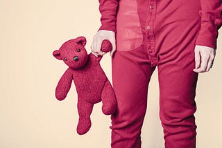 Image with a yellowish background, and to the right of the image stands a grown person wearing dark red and holding a teddy in the same color.