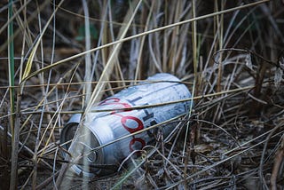 A discarded silver can of Coors Light beer lies on the ground amidst weeds and dirt.