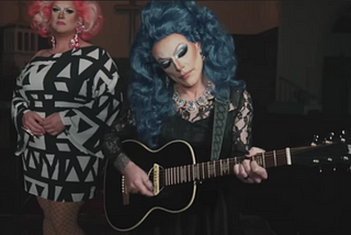 Two people in drag. The one in the back is wearing a black and white patterned dress and a pink wig. The one in the front is wearing a black lacy dress and a blue wig and playing a guitar.