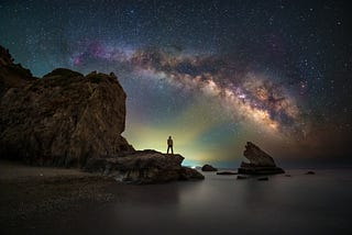 A man standing by the sea, with the large expanse of the sky above him and what appears to be Northern Lights.