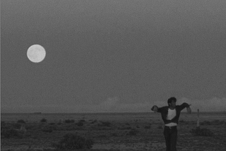 Screen shot from Badlands 1973, by Terrence Malick. A man stands in the desert at night with his arms draped lazily over a stick. The moon behind him is full and the subtitle reads: We lived in utter loneliness, neither here nor there.