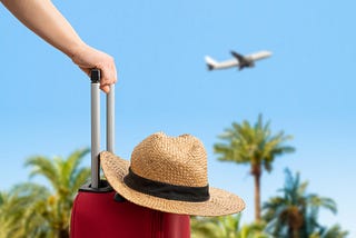 A hand on the handle of red suitcase with a hat on top and in the background, an airplane in the air.
