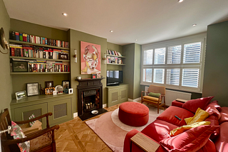 A photo of the author’s renovated living room which has olive green walls, a cherry red sofa, a pale pink rug and lots of books.