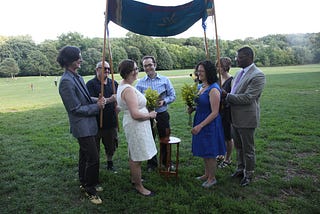 Author (in an off-white dress) and her wife at their Jewishly-inflected civil marriage ceremony, accompanied by their officiant, Cantor Joshua Breitzer, and their witnesses. Their chuppah is a Jewish prayer-shawl custom designed by Rabbi Nikki with textile artist Sharon Landecker.