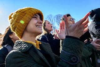 a person in a yellow knit hat, facing right, smiling and clapping, amid a crowd of people