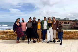 A group of Black trans and gender-expansive folks smiling and standing on payment close to a shore with a low dark yellow brick wall.