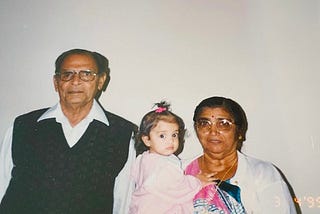 A baby, middle, wears a pink outfit and sits in the arms of her grandmother, a dark-skinned woman with a black bun and colorful saree. A man, the baby’s grandfather, stands to the left, wearing a white collared shirt and black vest.