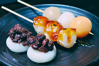 Three varieties of Japanese dango on a black plate. The front shows two white dango topped with sweet red bean paste (anko). In the middle, skewered dango are coated in a sweet soy sauce glaze (mitarashi). In the back, three-color dango in pink, white, and green are skewered.