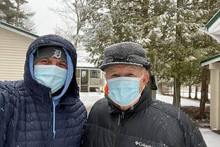 Father and son stand in front of the cabin while it’s snowing dressed in winter coats, hats, and surgical masks.