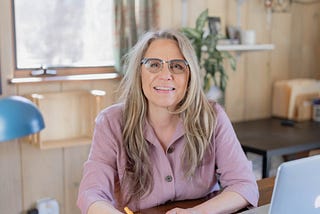 Photo of the author, Uma Bode, sitting at her desk facing the camera, smiling, holding a pencil in her hand. The background is out of focus and shows a window, a plant, and shelves.