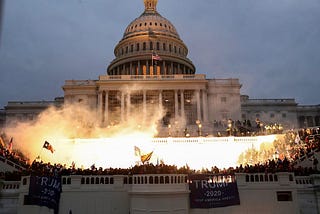 A photo of smoke in front of the U.S. Capitol Building; Trump 2020 flags can be seen hanging in front of a crowd of rioters.
