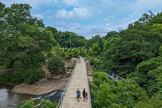 Pedestrians walking over a bridge crossing a river and bordered by green trees