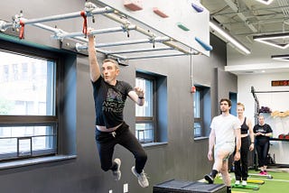 A man swings by one arm on monkey bars, wearing a black Amplify Fitness shirt and looking focused at the bar in front of him. Three people wait in the background for their turn.
