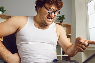 Man sitting in a white sleeveless undershirt and pj’s looks and points angrily at his computer.