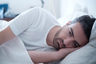The image is of a young man lying on his side in bed facing the camera, witha depressed look on his face.