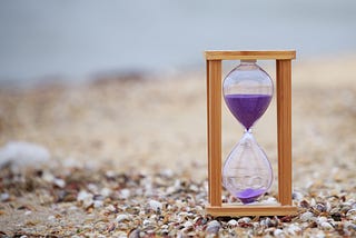 Close-up photo of a wooden hourglass with purple sand in it. Background is of a beach.