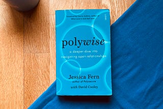 A paperback copy of “polywise: a deeper dive into navigating open relationships” by Jessica Fern with David Cooley. The book has a blue cover, and is resting on a wooden table with a folded blue tablecloth and a cup of coffee.