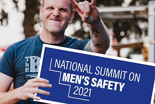 A young white man holds a sign for a Men’s Safety Summit while holding his hand up in the rock on gesture.