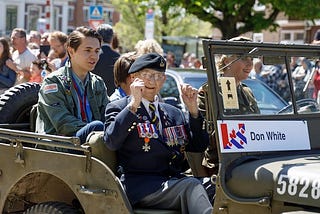 An old man with war medals waves from a jeep with a young citizen seated behind