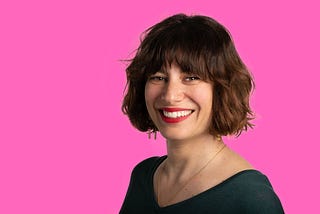 Photo of Anouk against a bright pink background, wearing red lipstick and smiling.