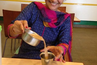 A woman pours filter coffee from a rimmed bowl to a rimmed glass. She is smiling. A bowl of something savory and yummy sits in front of her.