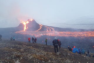Several spread out groups of people in winter clothes stand and sit, watching the eruption of a cinder cone volcano in Iceland in 2021.