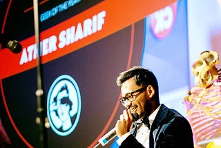 Ather holding a microphone on a stage with a big screen and a skeleton behind him.