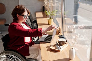 A woman with wheelchair facing her laptop in a table near big glass window