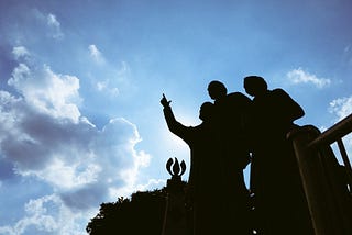An image of the Gateway to Freedom International Memorial in Detroit. I took this image from below so the blue sky is visible and the memorial is shadowed. We see a person standing with others and pointing forward.