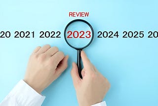 A picture of a pair of hands rested on a blue board. On the blue board are the words Review, 2020, 2021, 2022, 2023, 2024, and 2025. The word “Review” and “2023” are in red. One hand is holding a magnifying glass over the year 2023.